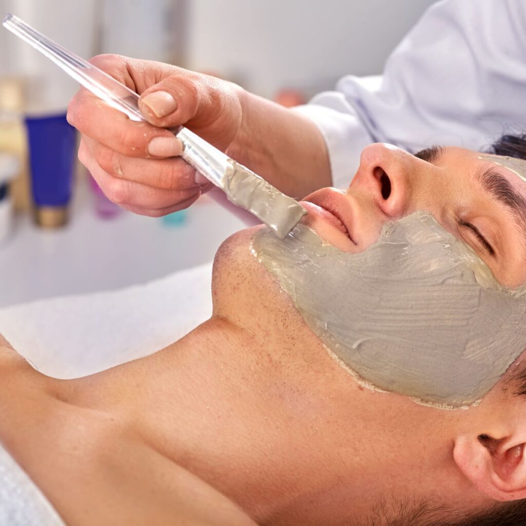 Man at a spa gets a facial from an esthetician, who hopefully practices infection control techniques by cleaning the brush in hand after every client’s face mask.