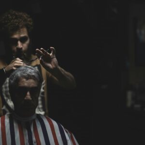 Barber cuts a man’s hair in a dark, ominous space, as if remedying barber shop complaints.