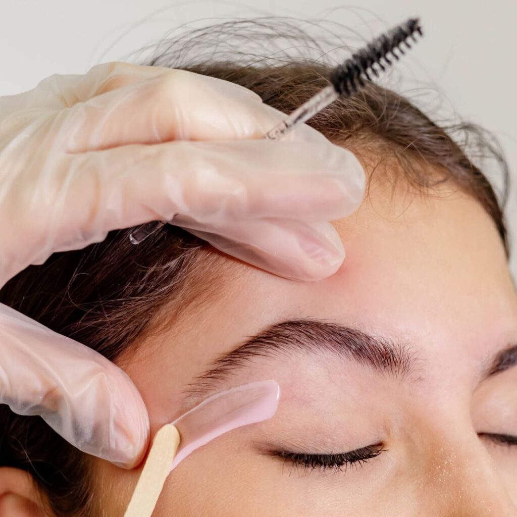 Waxing specialist wearing gloves for waxing applies treatment under a woman's eyebrows with a single-use waxing popsicle stick. Proper technique can reduce your risk of waxing dangers.