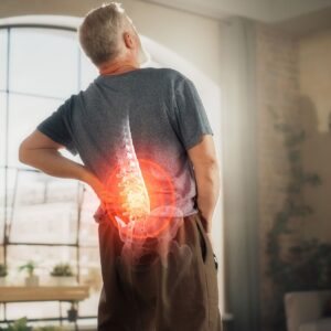 Man standing in an apartment holding his lower back in pain, with a circle around his lower spine indicating inflammation and discomfort after an injury from massage.