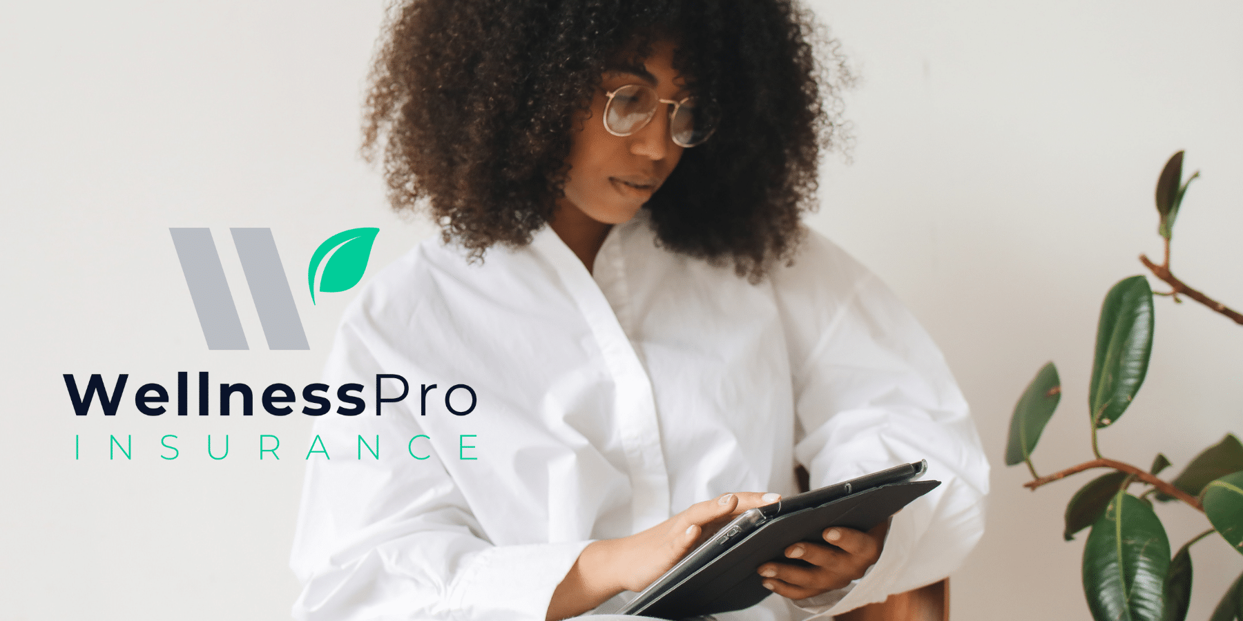 Woman wearing glasses sits next to a green plant while looking down at a grey tablet, as if reading small business insurance tips and advice on the WellnessPro insurance blog.