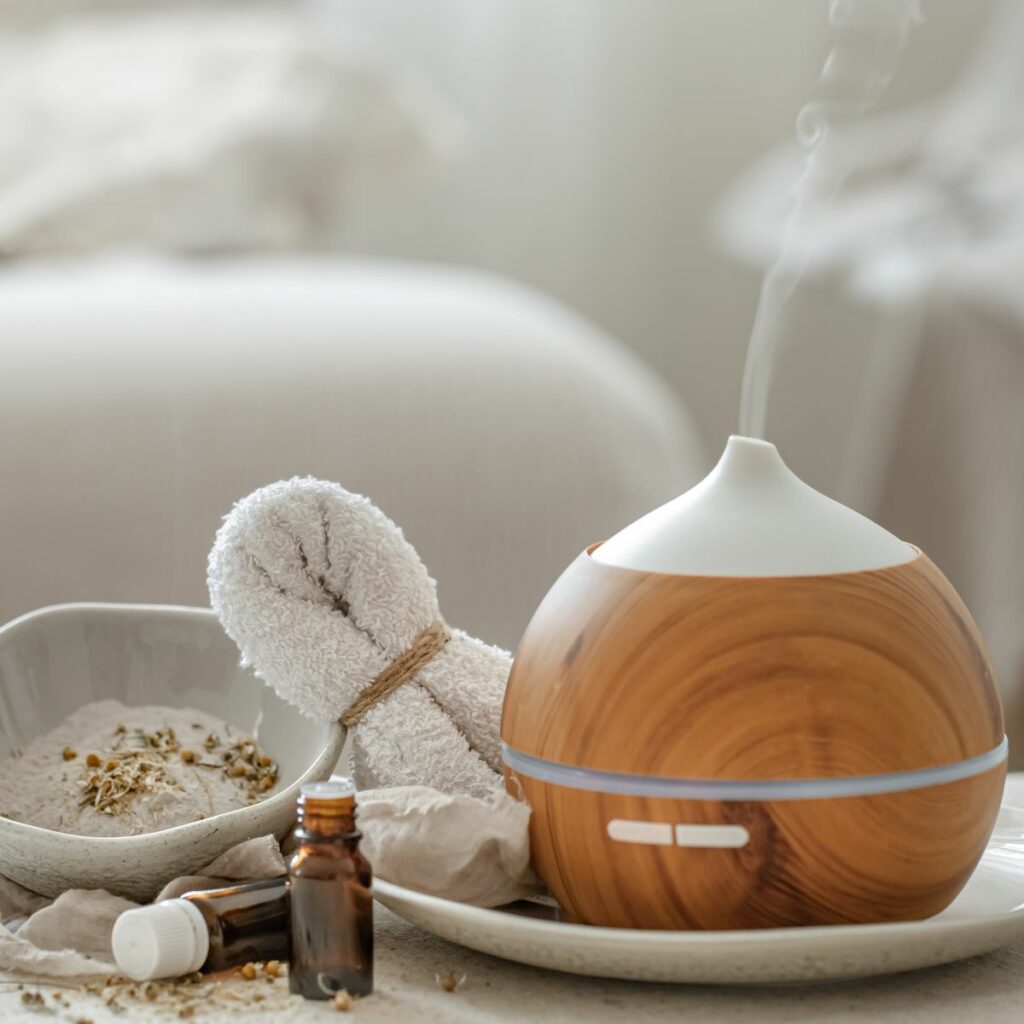 Essential oil diffuser displayed in a wellness spa next to bottles of essential oils, a bowl of dried plants, and a washcloth. Massage aromatherapy services like this one should address health claims regarding essential oils to prevent accidents.