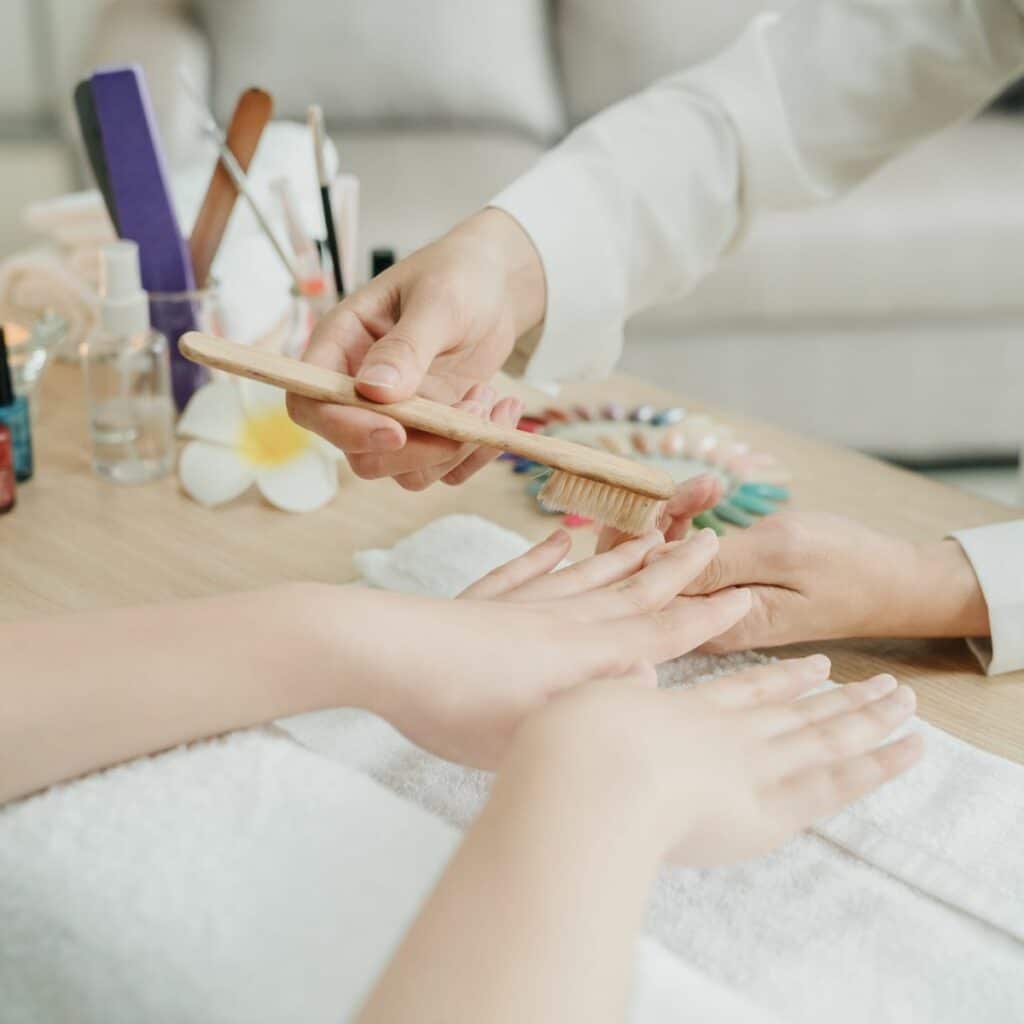 Image of a home nail salon setup, featuring a nail tech brushing their client’s nails with nail polish bottles, candles, and a couch off to the side. Nail tech displays professionalism needed for a nail studio at home.