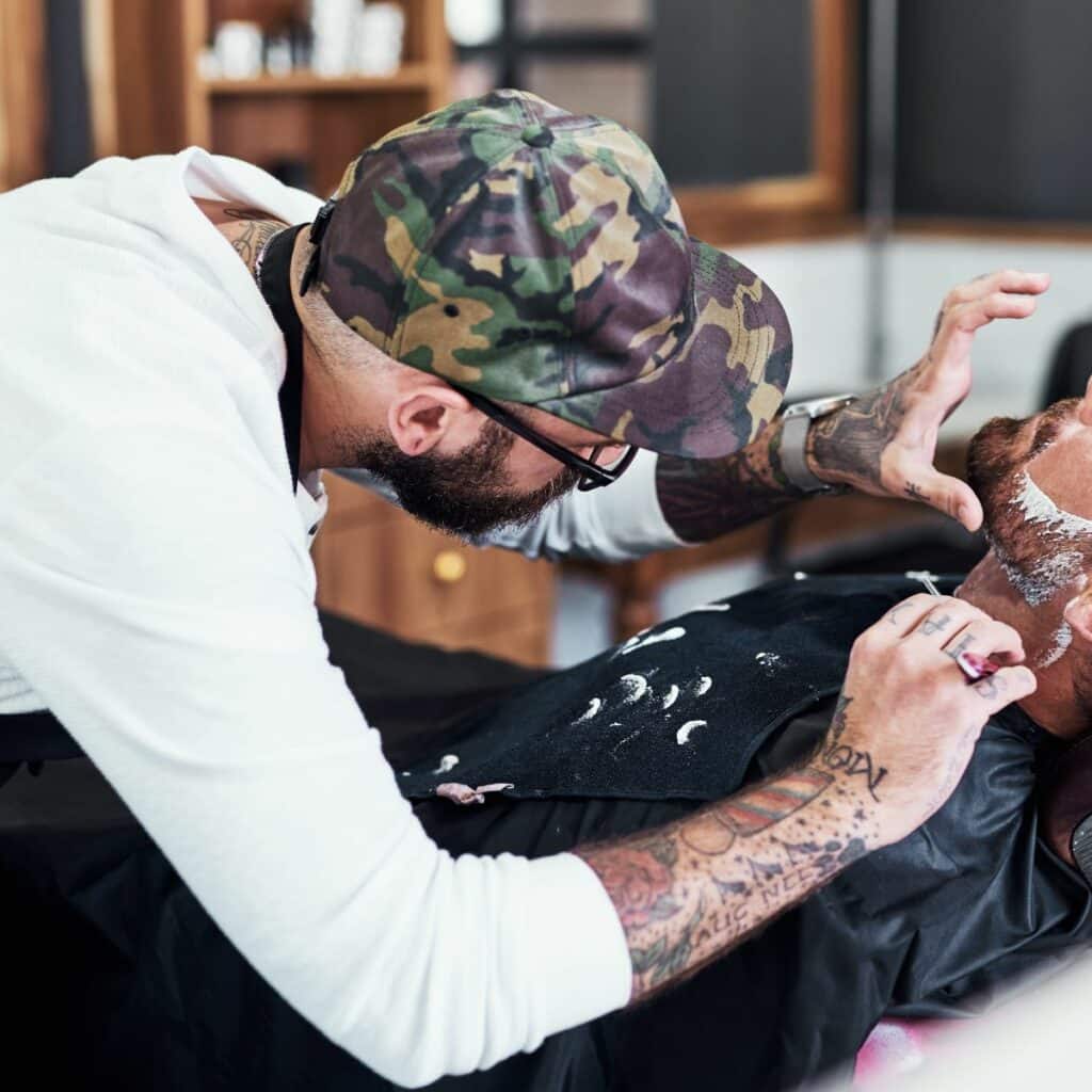 Barber wearing a baseball hat and glasses carefully avoids nicks and cuts on a client’s face while shaving his beard. By gently pulling the reclined man’s skin on sensitive areas like the neck, he’s less likely to cause a barber shop razor cut.