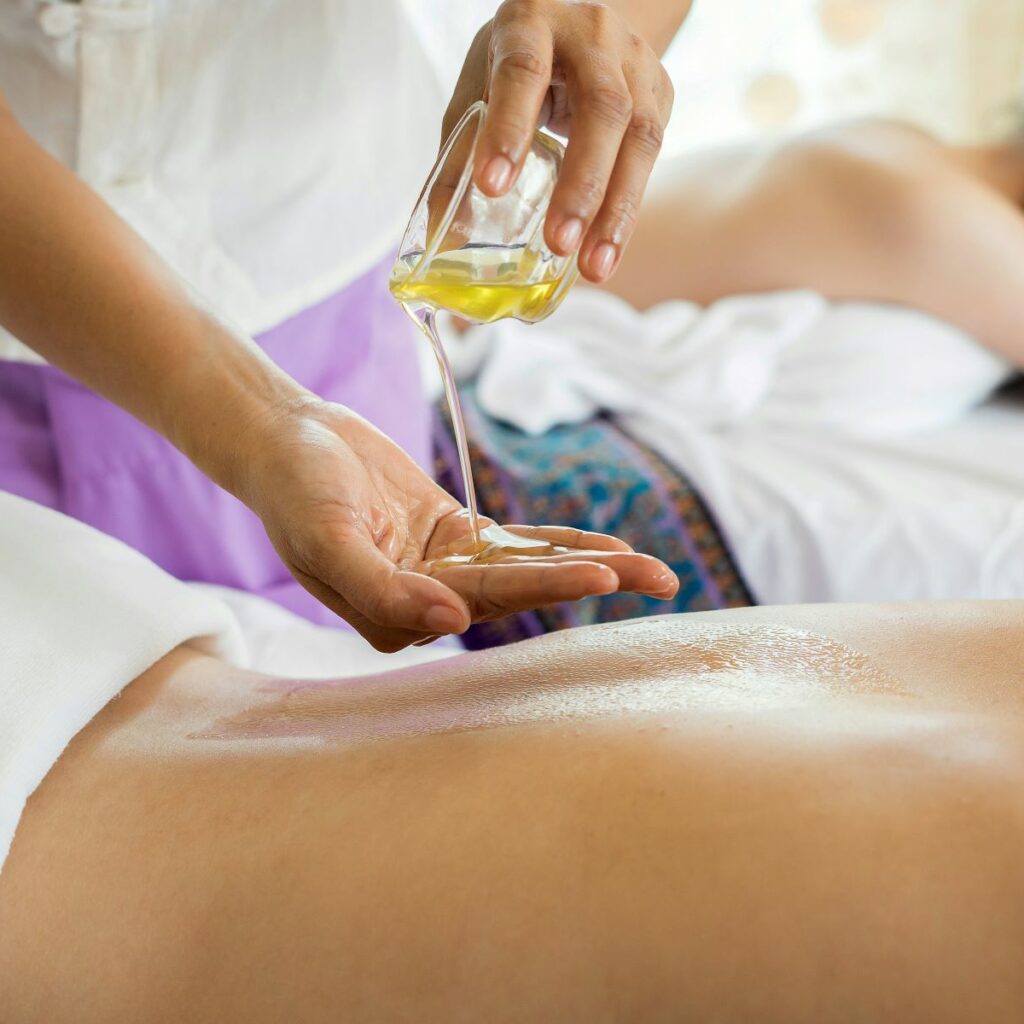 Two massage therapy clients lying on massage tables while their masseuse pours oil. Clients and masseuse appear relaxed, unlike those who are stressing over the question, “Do massage therapists need insurance”?