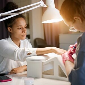 Client sits at a workstation while her nail technician uses an electric filer on her nails. The tech puts the client’s arm and wrist on an elevated surface, an ergonomics best practice for preventing nail technician pain, a repetitive strain injury for nail technicians, like carpal tunnel syndrome, and other nail tech problems.