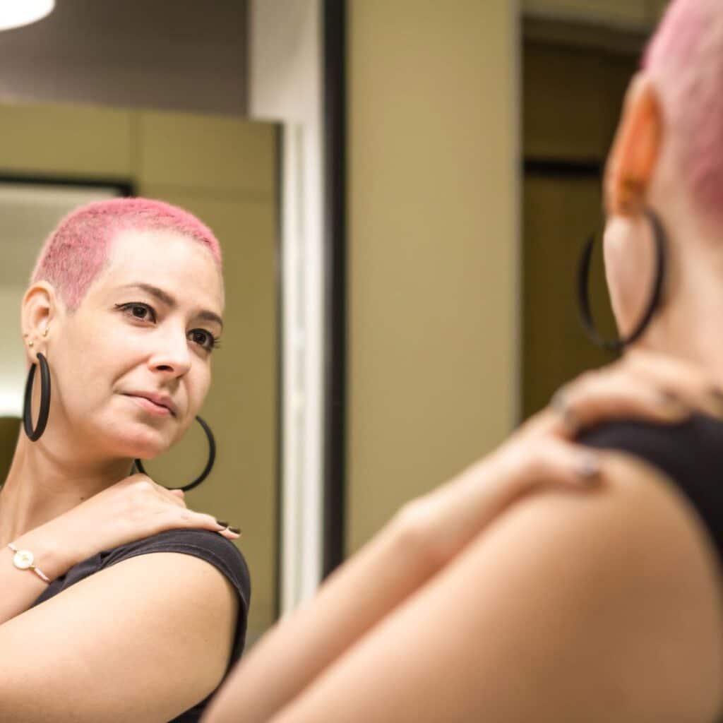 Person with short, pink hair stands strong and confident while observing themself in the mirror, presumably after finishing chemotherapy. Hair plays a key role in body positivity and self-image, leading many to seek hair coloring after chemotherapy or even dying hair while on chemo.