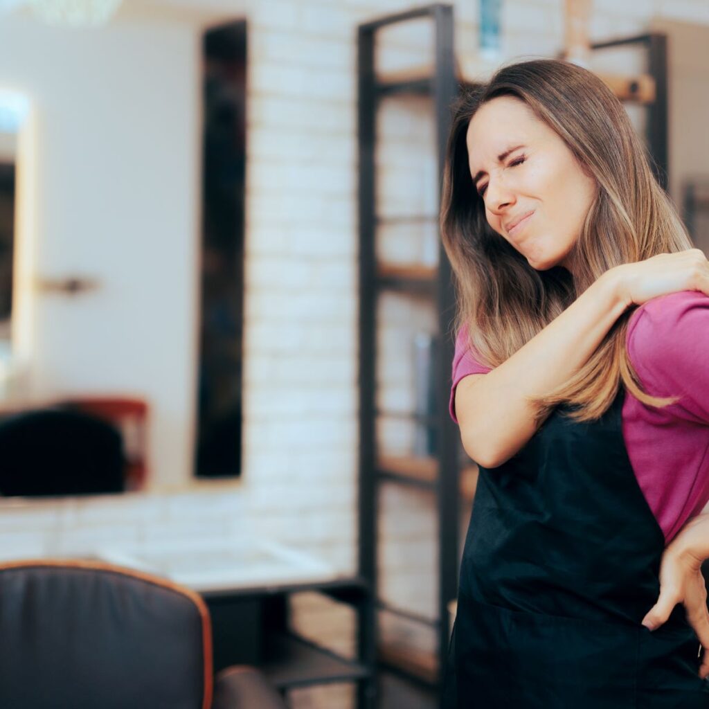 A hair salon stylist grimaces in pain from possible shoulder overuse injuries. Hairdresser shoulder pain and back pain can be prevented with stretches for hairdressers.