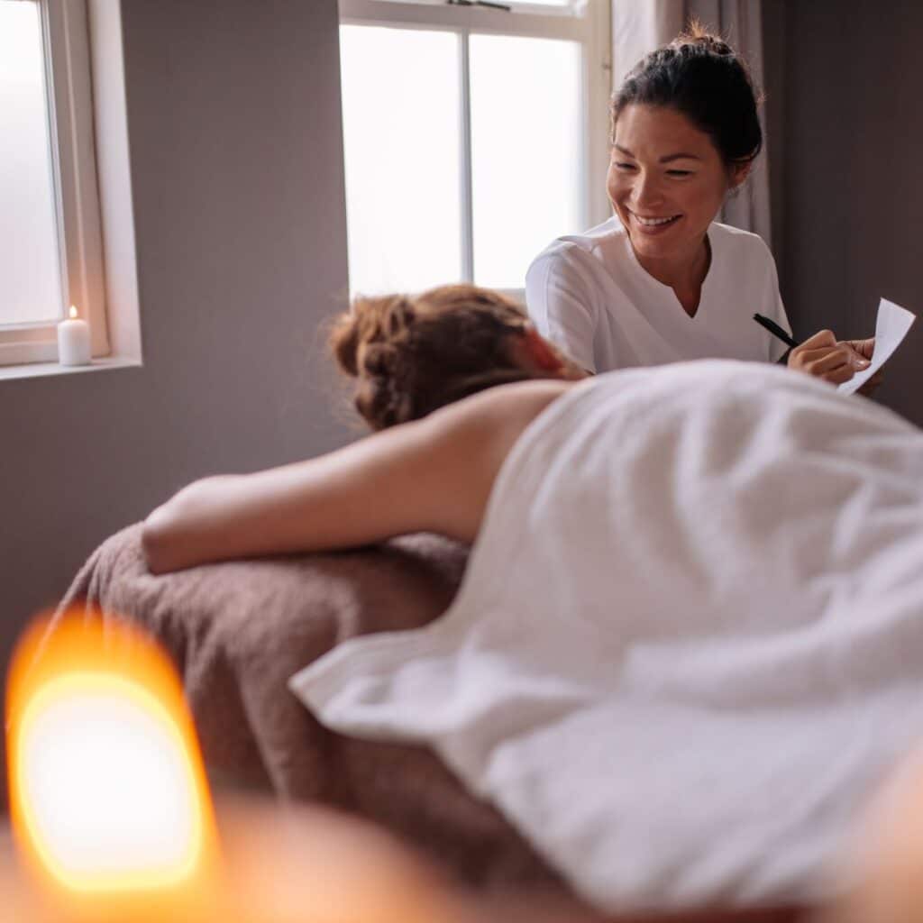 A massage therapist smiles at a client getting a massage for the first time. She takes notes on paper and chats with the first-time massage client about how to make the massage client more comfortable.