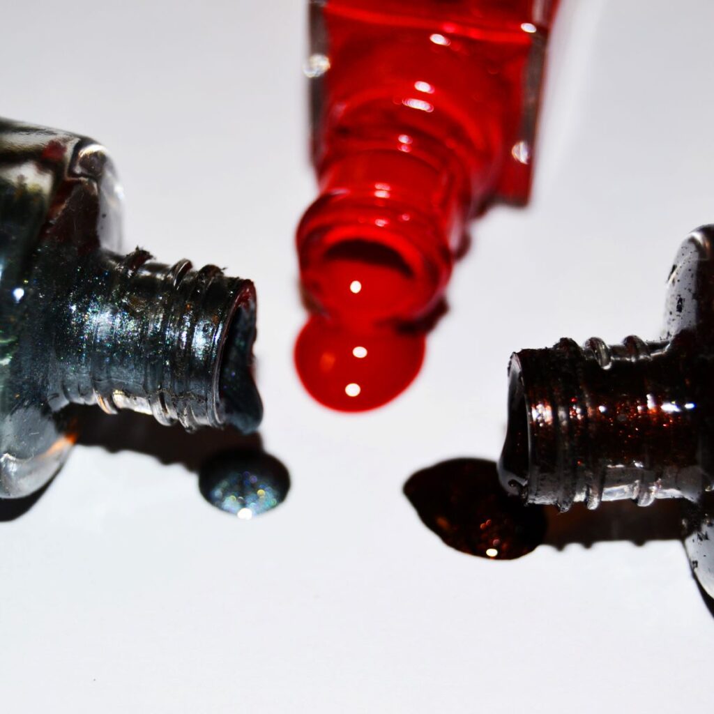 Three bottles of dark-colored nail polish tipped over and spilled onto a white surface, alluding to hazardous chemicals found in nail salon products and ingredients in nail polish.