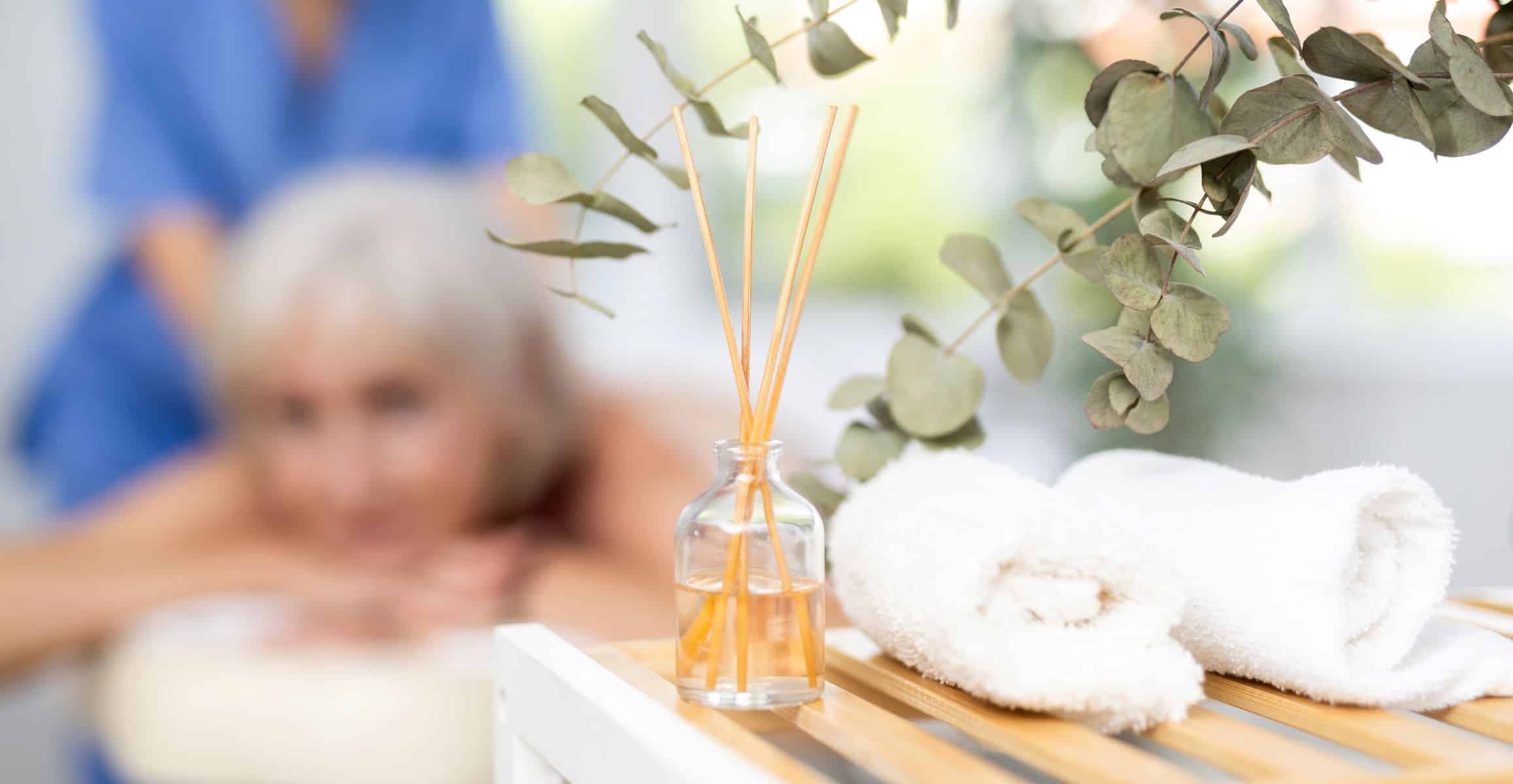 Client lying on a massage table in the background out of focus, with a table of odor eliminating essential oils rods and a eucalyptus plant in focus in the foreground.