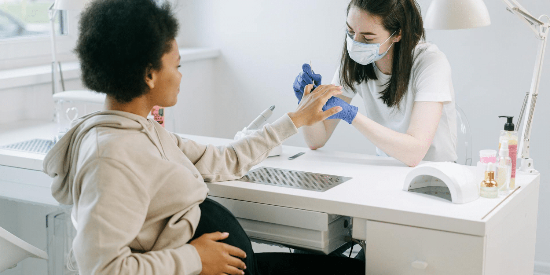 Nail artist wearing protective gloves and a mask pushes back cuticles of pregnant client in the salon chair.