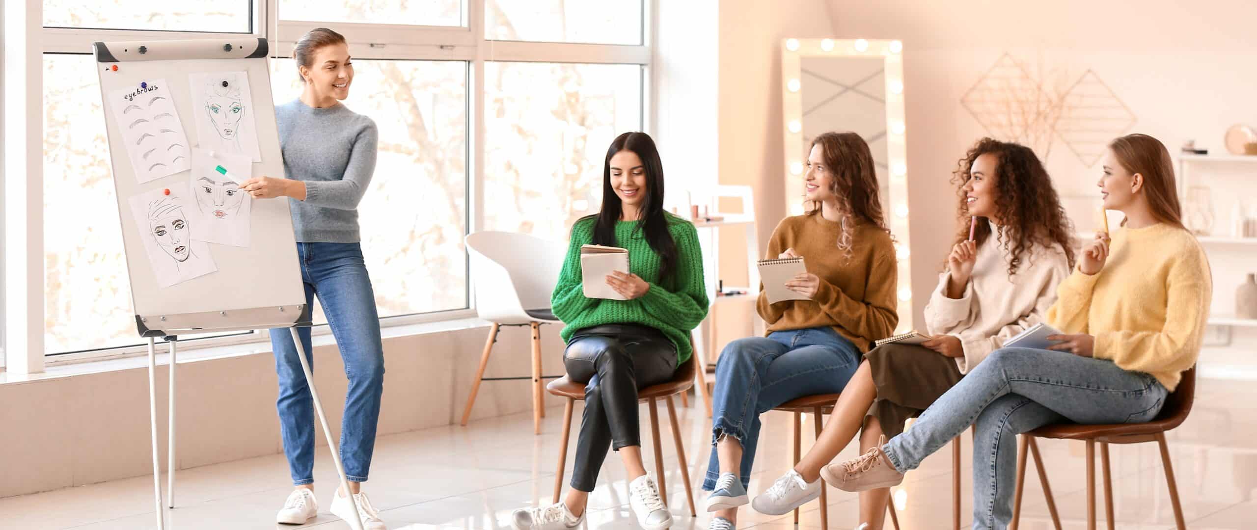 Four women sit in chairs next to a teacher who uses illustrations to teach about skincare or makeup, like esthetician continuing education classes.