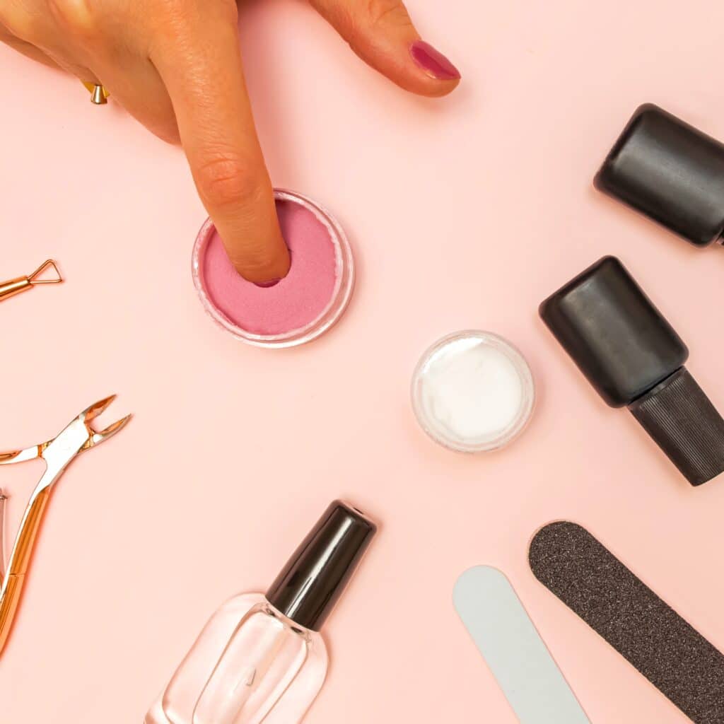Nail technician’s client dips their finger into pink nail powder, likely having already taken steps to prevent side effects of dip powder nails using surrounding tools (nail file, cuticle trimmer, bottles) safely.