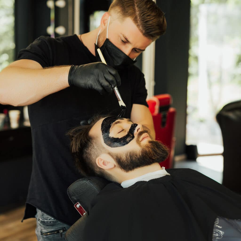 Seated male client gets a clarifying face mask as part of today’s luxury men's grooming trends and premium beard grooming services.