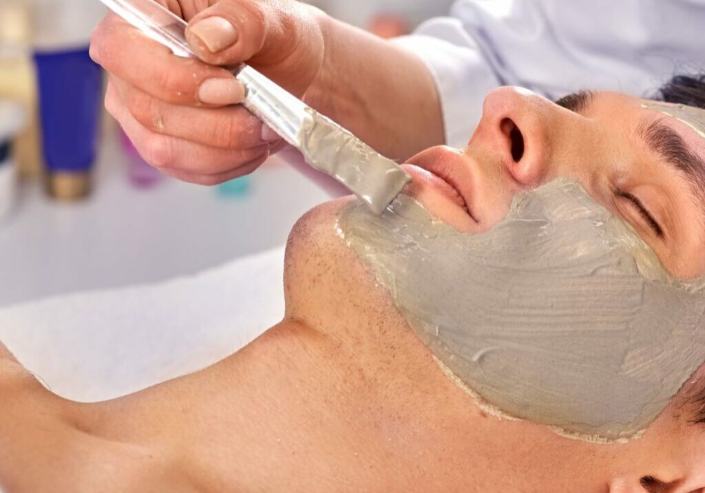 Man at a spa gets a facial from an esthetician, who hopefully practices infection control techniques by cleaning the brush in hand after every client’s face mask.