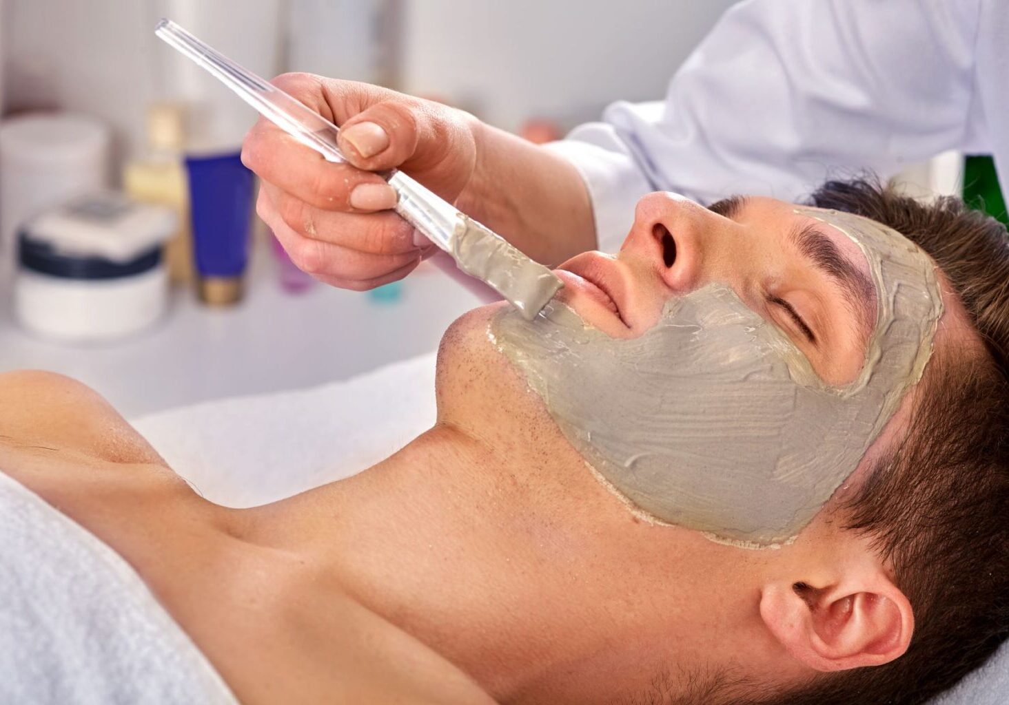 Man at a spa gets a facial from an esthetician, who hopefully practices infection control techniques by cleaning the brush in hand after every client’s face mask.