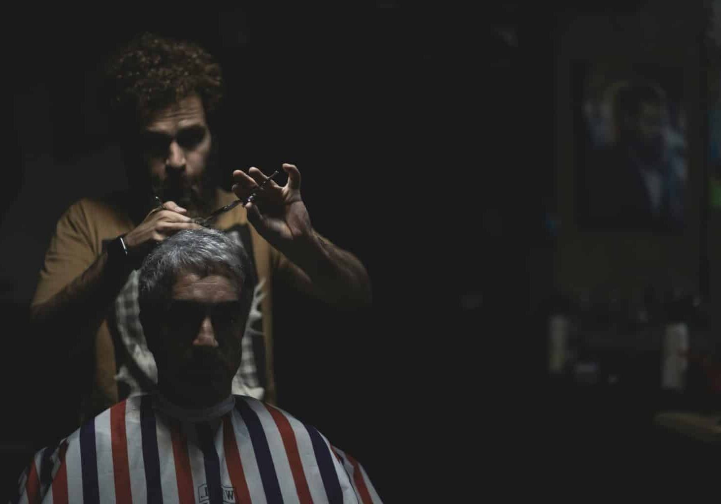 Barber cuts a man’s hair in a dark, ominous space, as if remedying barber shop complaints.