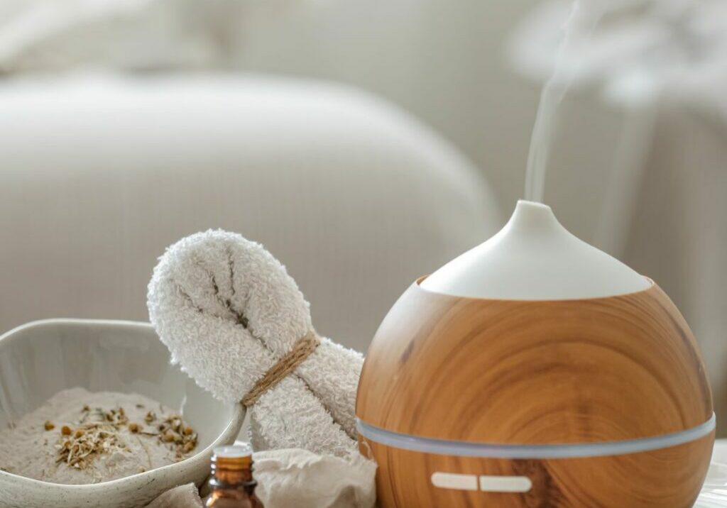 Essential oil diffuser displayed in a wellness spa next to bottles of essential oils, a bowl of dried plants, and a washcloth. Massage aromatherapy services like this one should address health claims regarding essential oils to prevent accidents.