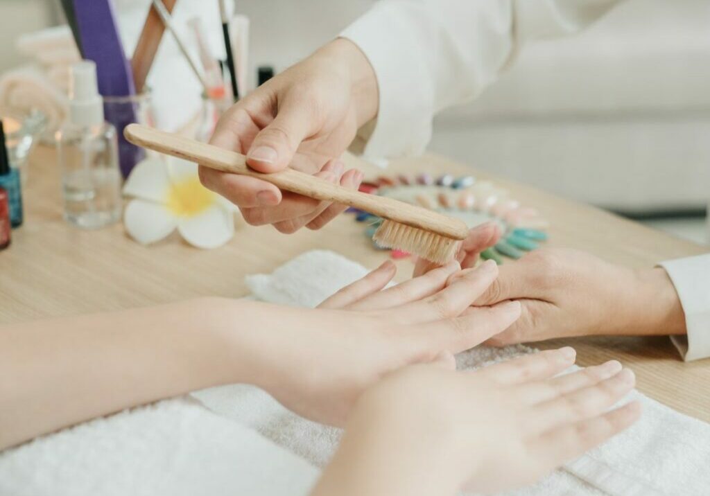 Image of a home nail salon setup, featuring a nail tech brushing their client’s nails with nail polish bottles, candles, and a couch off to the side. Nail tech displays professionalism needed for a nail studio at home.