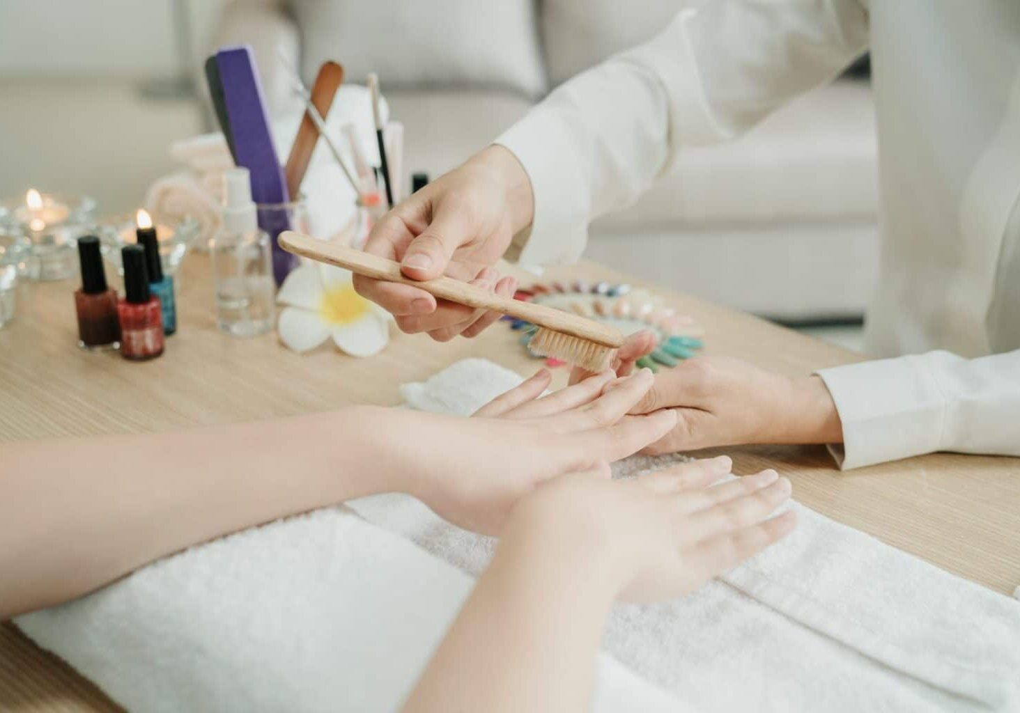 Image of a home nail salon setup, featuring a nail tech brushing their client’s nails with nail polish bottles, candles, and a couch off to the side. Nail tech displays professionalism needed for a nail studio at home.