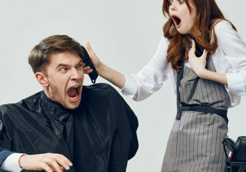 Man in a barbershop or salon chair with a shocked expression while his barber or hairstylist seems to accidentally cut him with clippers. A bad cut could lead to a hair salon lawsuit or barber problem.