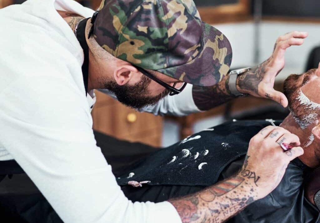 Barber wearing a baseball hat and glasses carefully avoids nicks and cuts on a client’s face while shaving his beard. By gently pulling the reclined man’s skin on sensitive areas like the neck, he’s less likely to cause a barber shop razor cut.