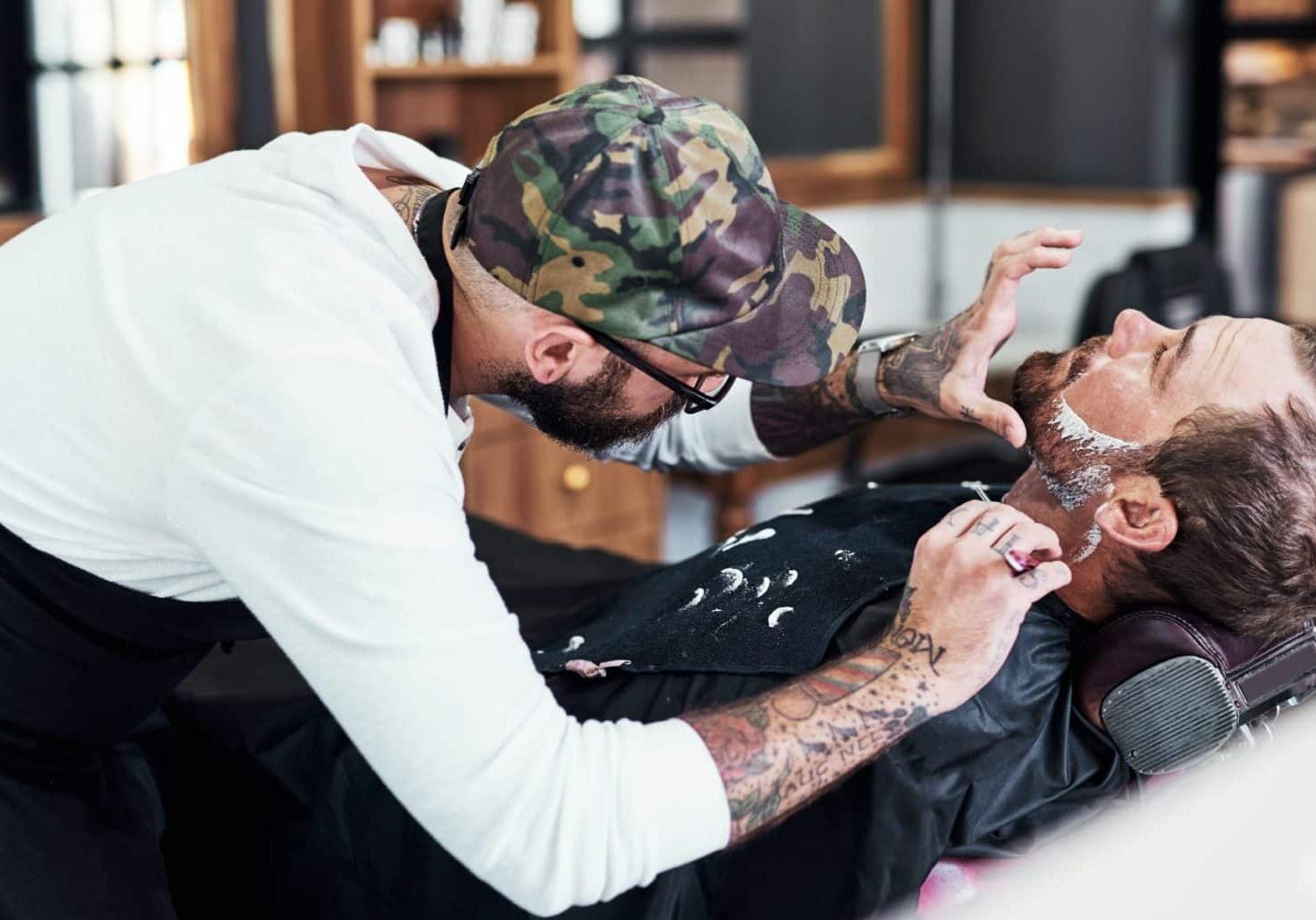 Barber wearing a baseball hat and glasses carefully avoids nicks and cuts on a client’s face while shaving his beard. By gently pulling the reclined man’s skin on sensitive areas like the neck, he’s less likely to cause a barber shop razor cut.