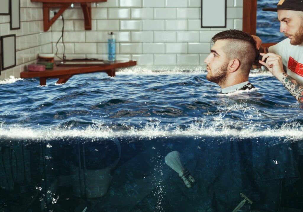 Man seated in a barbershop chair is getting his hair cut by a barber. Meanwhile, they’re surrounded by water from a flooded salon. A barbershop with this much standing water would face serious mold growth signs of water damage in the walls.