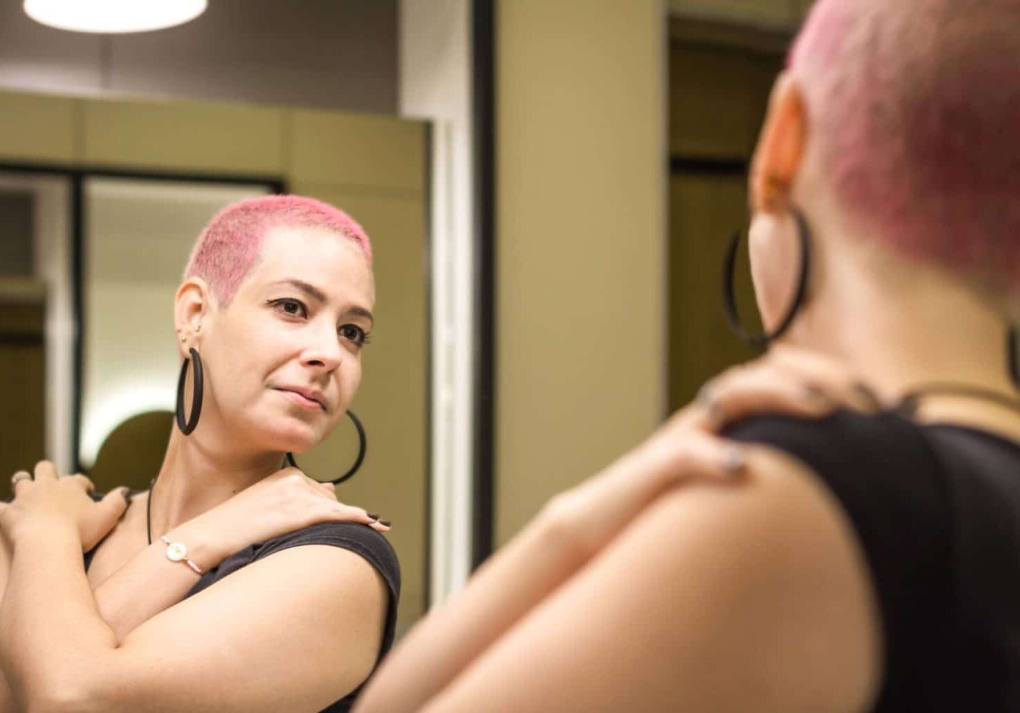 Person with short, pink hair stands strong and confident while observing themself in the mirror, presumably after finishing chemotherapy. Hair plays a key role in body positivity and self-image, leading many to seek hair coloring after chemotherapy or even dying hair while on chemo.