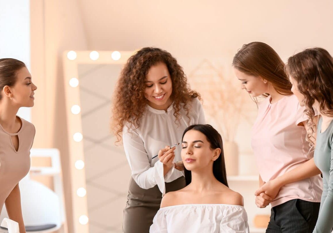 continuing-education-for-estheticians Cosmetology students gather around a teacher demonstrating how to apply product on a model’s face, likely skincare specialist courses or a class for continuing education for estheticians.
