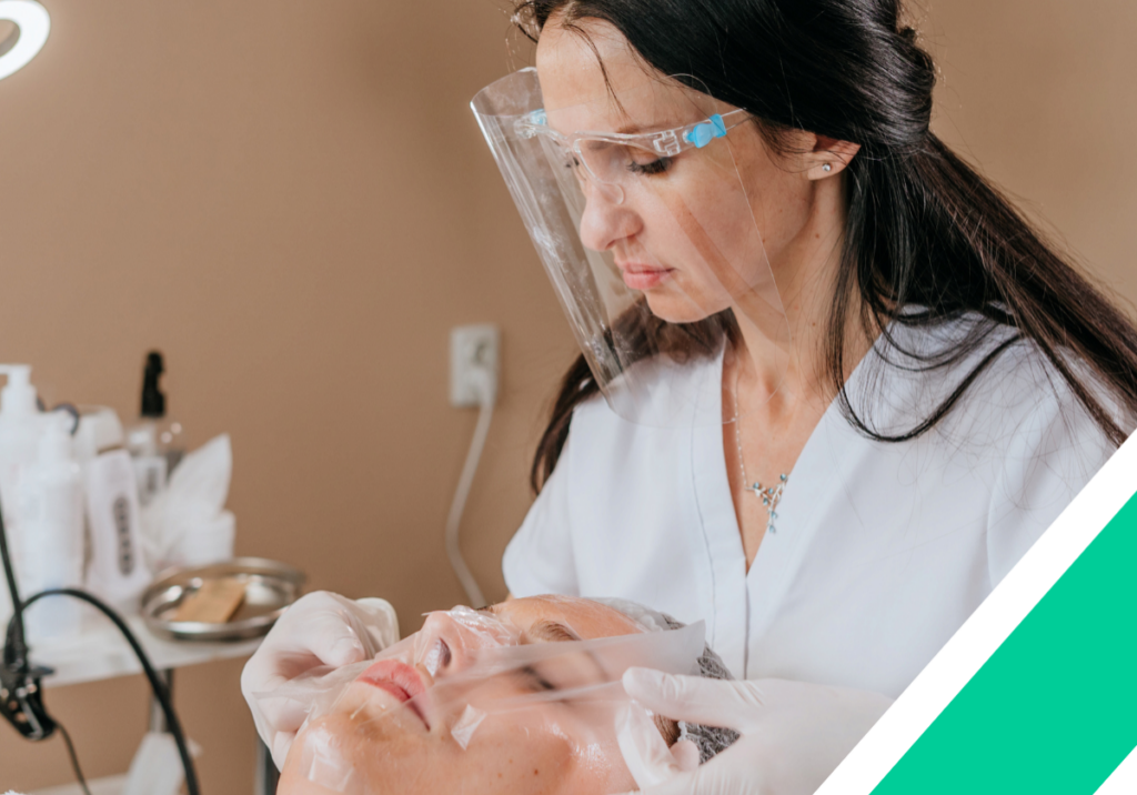 Esthetician or aesthetician wearing a clear plastic face mask puts paper on client’s face to perform a skin specialist procedure.