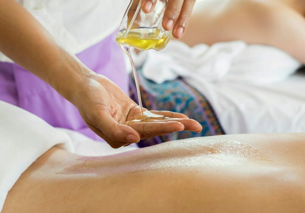 Two massage therapy clients lying on massage tables while their masseuse pours oil. Clients and masseuse appear relaxed, unlike those who are stressing over the question, “Do massage therapists need insurance”?