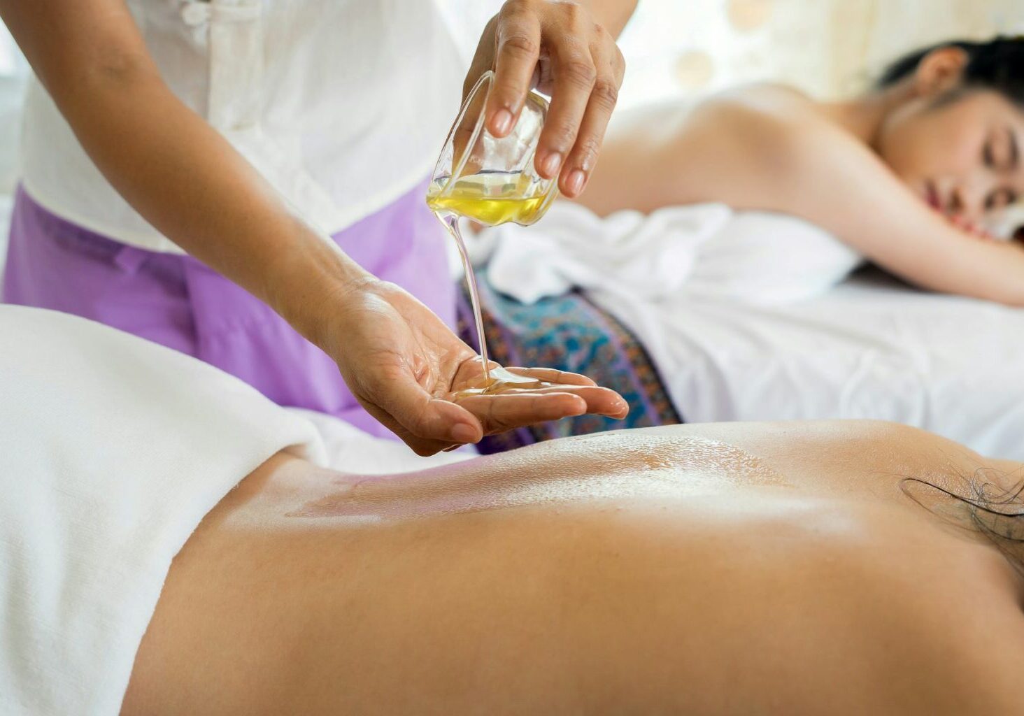 Two massage therapy clients lying on massage tables while their masseuse pours oil. Clients and masseuse appear relaxed, unlike those who are stressing over the question, “Do massage therapists need insurance”?