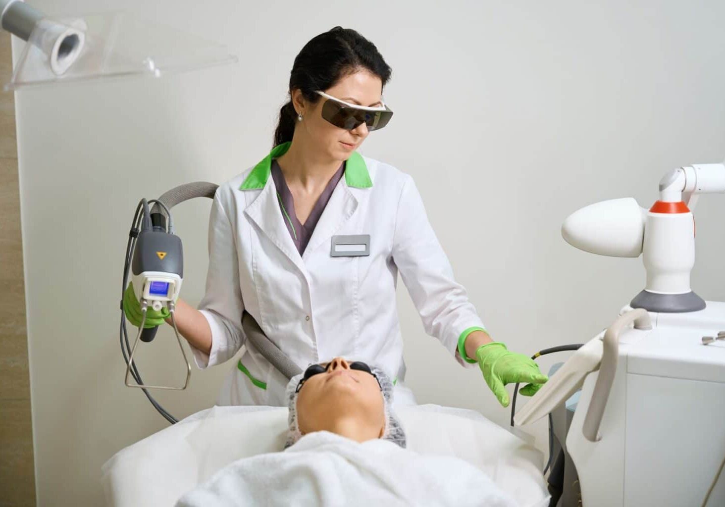 While working as an esthetician, a woman wearing safety goggles, protective gloves, and a lab coat demonstrates a safe esthetician work environment while performing a laser procedure on a client.