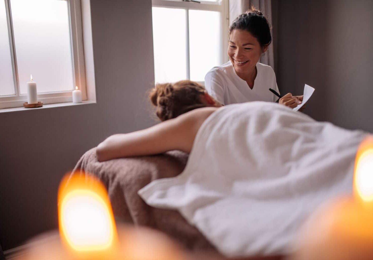 A massage therapist smiles at a client getting a massage for the first time. She takes notes on paper and chats with the first-time massage client about how to make the massage client more comfortable.