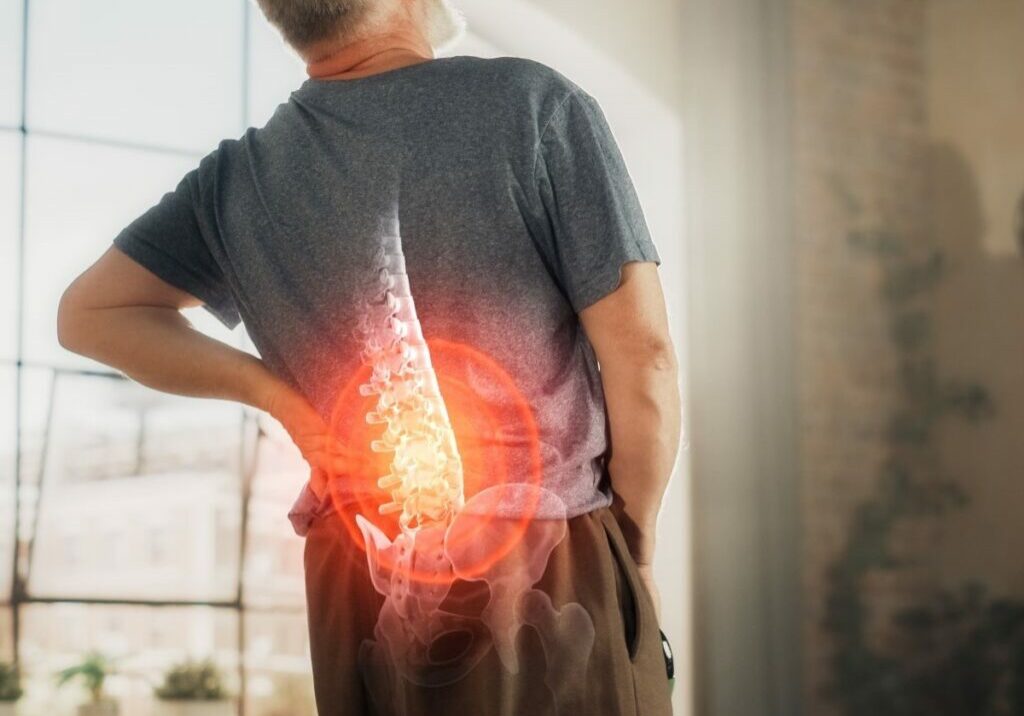 Man standing in an apartment holding his lower back in pain, with a circle around his lower spine indicating inflammation and discomfort after an injury from massage.