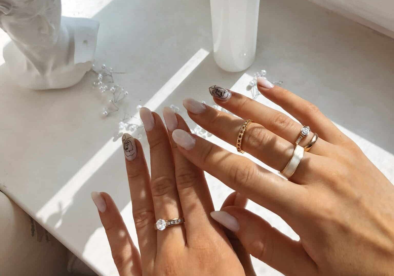 nail-enhancements Woman’s hands wearing assorted rings on a marble counter next to a miniature bust statue, with different types of nail enhancements painted on her fingernails.