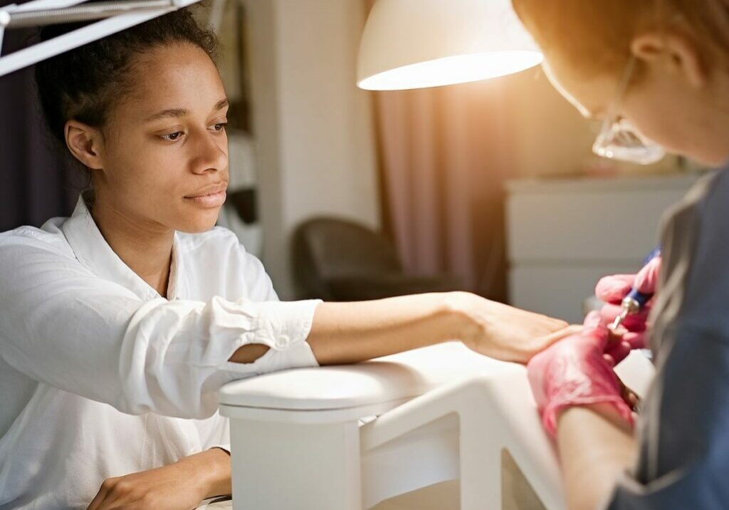 Client sits at a workstation while her nail technician uses an electric filer on her nails. The tech puts the client’s arm and wrist on an elevated surface, an ergonomics best practice for preventing nail technician pain, a repetitive strain injury for nail technicians, like carpal tunnel syndrome, and other nail tech problems.