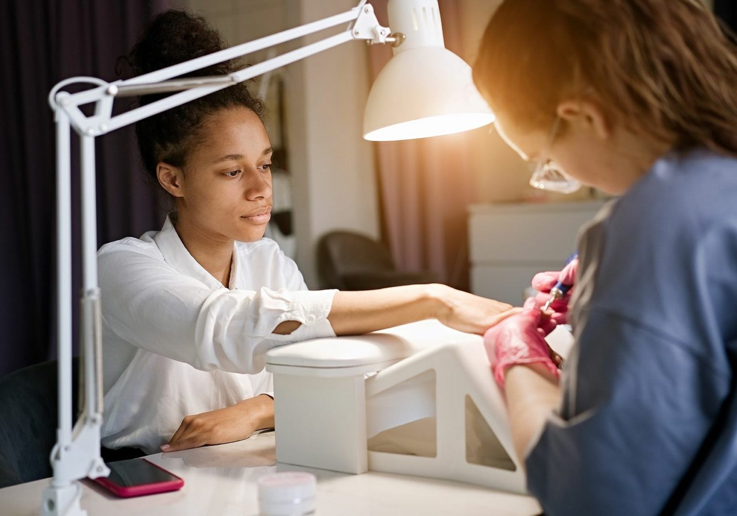 Client sits at a workstation while her nail technician uses an electric filer on her nails. The tech puts the client’s arm and wrist on an elevated surface, an ergonomics best practice for preventing nail technician pain, a repetitive strain injury for nail technicians, like carpal tunnel syndrome, and other nail tech problems.