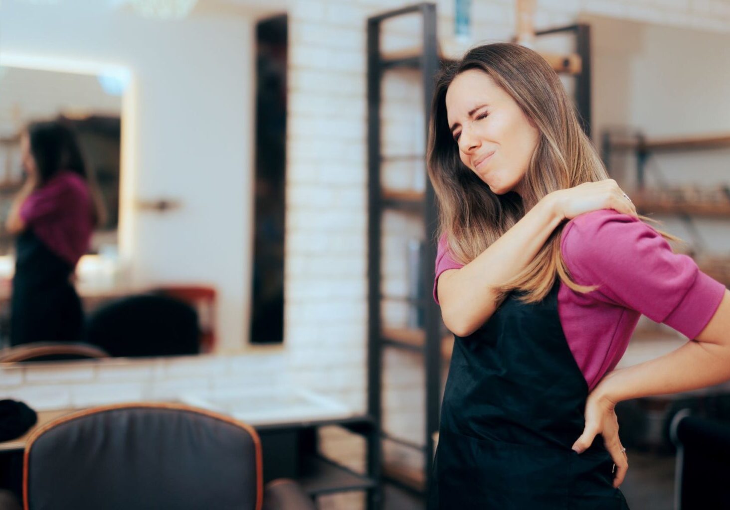 A hair salon stylist grimaces in pain from possible shoulder overuse injuries. Hairdresser shoulder pain and back pain can be prevented with stretches for hairdressers.