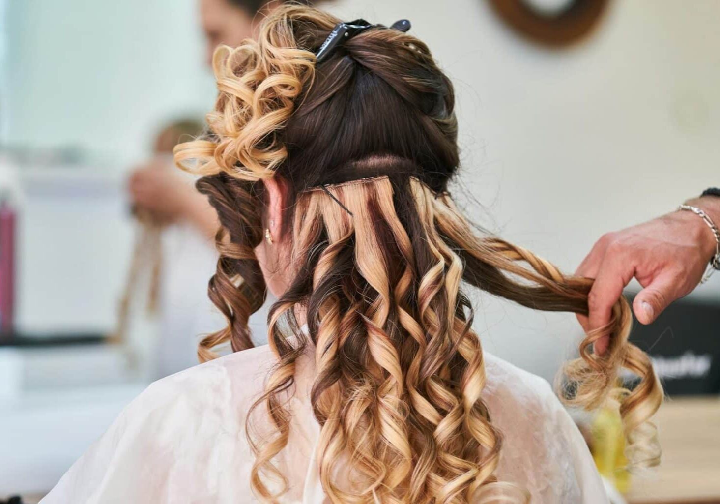 preventing-hair-extensions-damage Back of a client’s head while hairstylist installs hair extensions and curls the hair, likely working to prevent hair extensions damage and side effects of hair extensions.