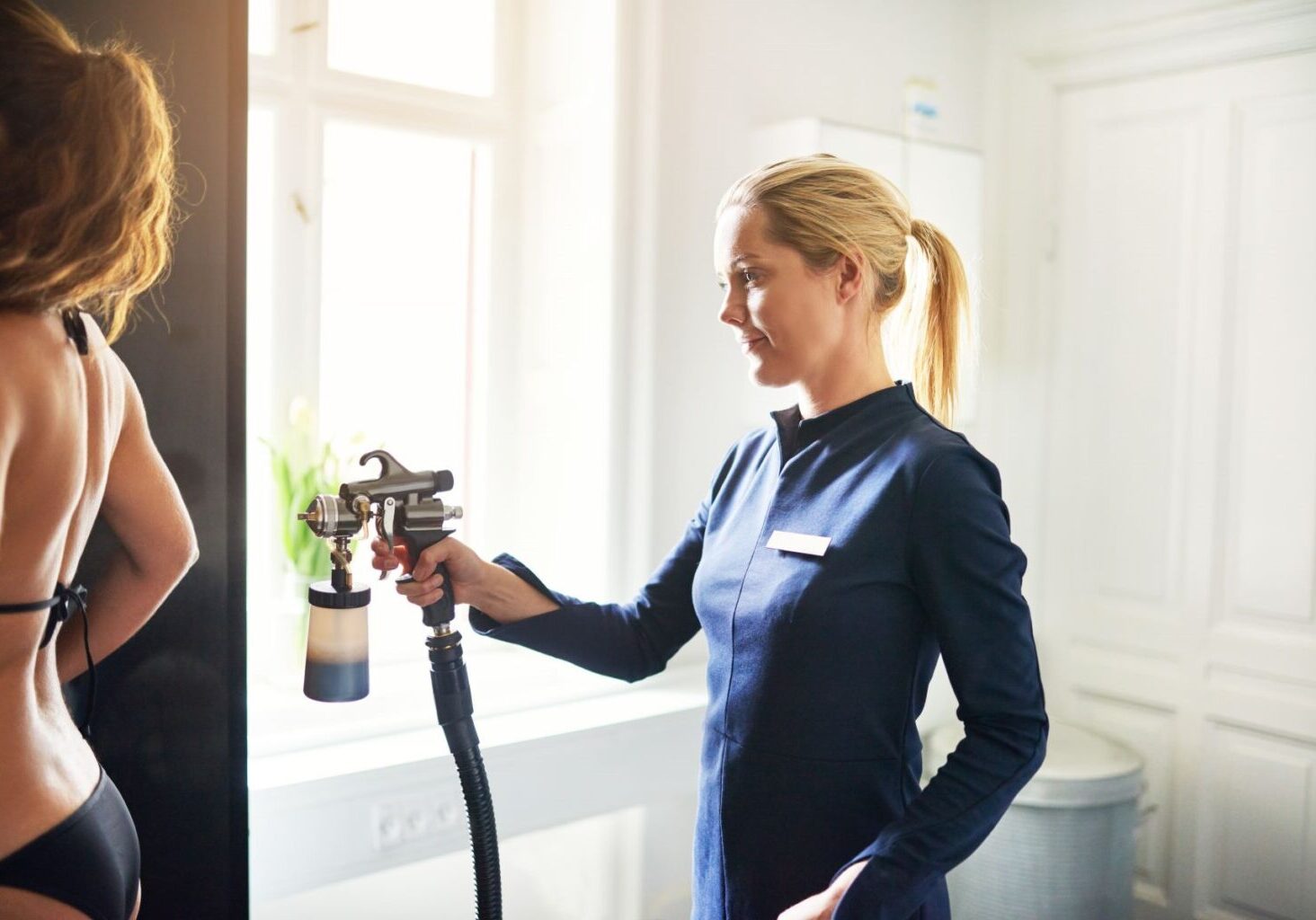 Female spray tan specialist wearing a blue uniform and using a spray bottle on a female client. Specialists like this should follow spray tan tips like standing farther away from clients.