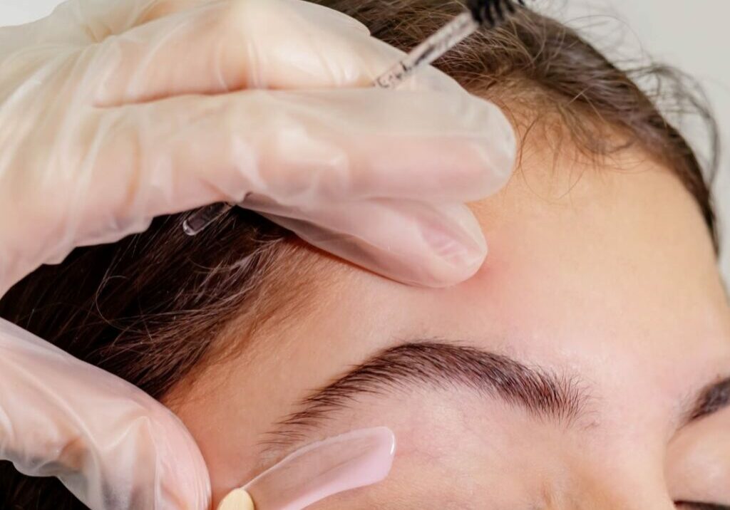 Waxing specialist wearing gloves for waxing applies treatment under a woman's eyebrows with a single-use waxing popsicle stick. Proper technique can reduce your risk of waxing dangers.