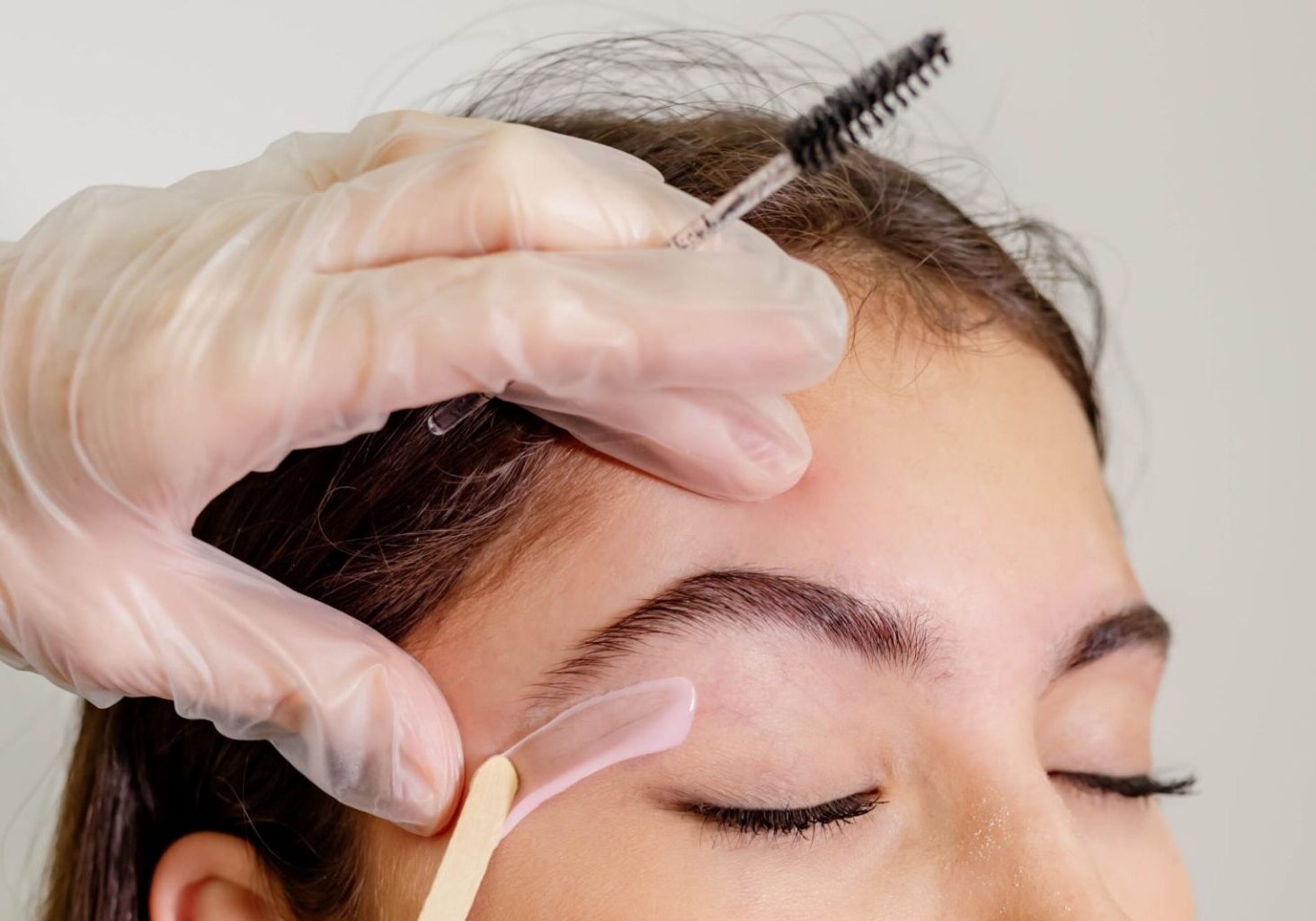 Waxing specialist wearing gloves for waxing applies treatment under a woman's eyebrows with a single-use waxing popsicle stick. Proper technique can reduce your risk of waxing dangers.